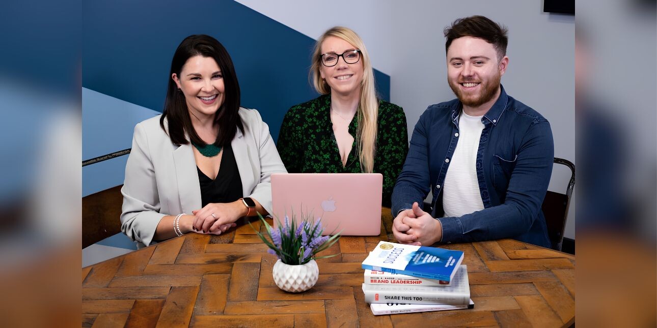 Three business colleagues sat around a table smiling at the camera: a white woman with dark hair, a white woman with blond hair (Samantha), and a man with dark hair. On the table are a pink laptop, plant and a pile of books. The background is blue an